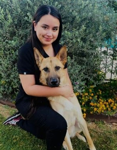 A woman kneels on the grass, hugging a large tan dog with pointy ears in a garden setting. Dressed in black, she resembles a caring veterinarian amid green bushes and yellow flowers, exuding warmth and affection for her furry friend.