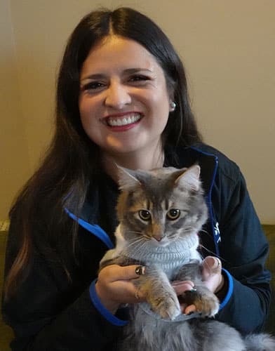 A smiling person with long dark hair holds a fluffy gray cat in a sweater, looking every bit like a caring veterinarian in their cozy, beige-walled home.