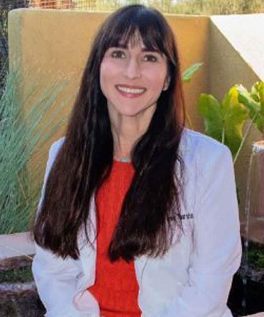 A smiling woman with long dark hair, clad in a white coat over a red top, sits outdoors amid greenery and a yellow wall. Her caring demeanor hints at her compassionate role as a veterinarian.