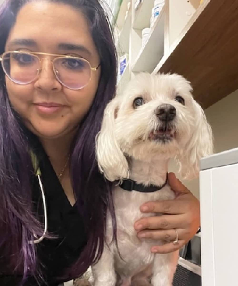 A woman with glasses and long dark hair, who could easily pass for a veterinarian, is taking a selfie with her small white dog wearing a black collar. She smiles warmly while holding the pup beside her in an indoor setting with shelves in the background.