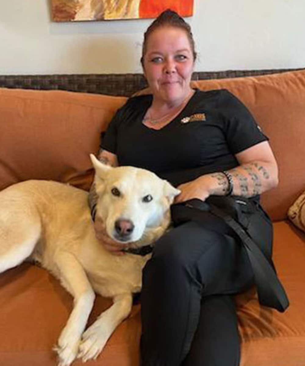 A woman with tattoos sits on a brown couch, smiling, and wearing black scrubs that mark her as a veterinarian. Her red hair is tied back. A white dog lies beside her with its head on her lap. A multi-colored abstract painting hangs on the wall behind them.