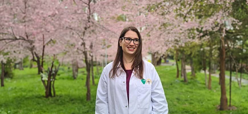 A veterinarian wearing glasses and a white coat stands smiling in a park filled with blossoming cherry trees. The green grass and light pink flowers create a serene, springtime atmosphere, as if nature herself is celebrating the care given to her creatures.