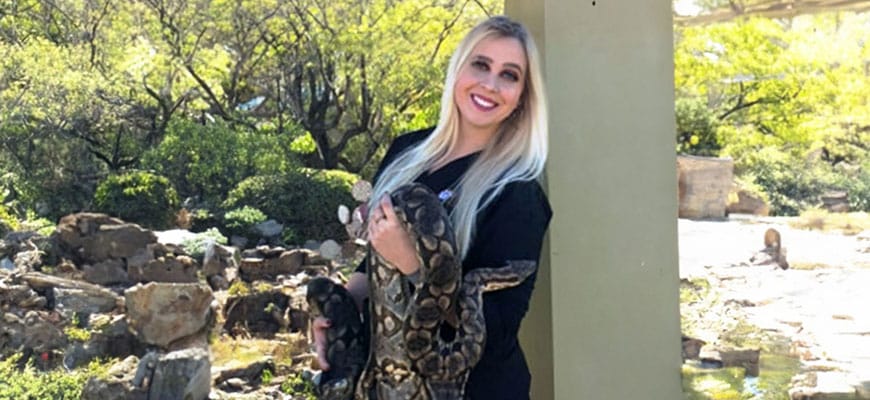 A woman with long blonde hair, wearing sunglasses and a black top, smiles while holding a large snake. As a veterinarian, she exudes confidence in the natural setting with trees and rocks in the background.