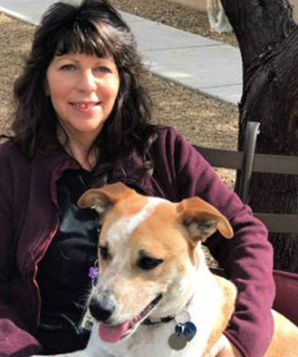 A woman with long dark hair is sitting outdoors on a bench, smiling at the camera. A brown and white dog, her companion from the veterinary clinic, rests in her lap. In the background, a tree trunk and a pathway add to the serene setting.