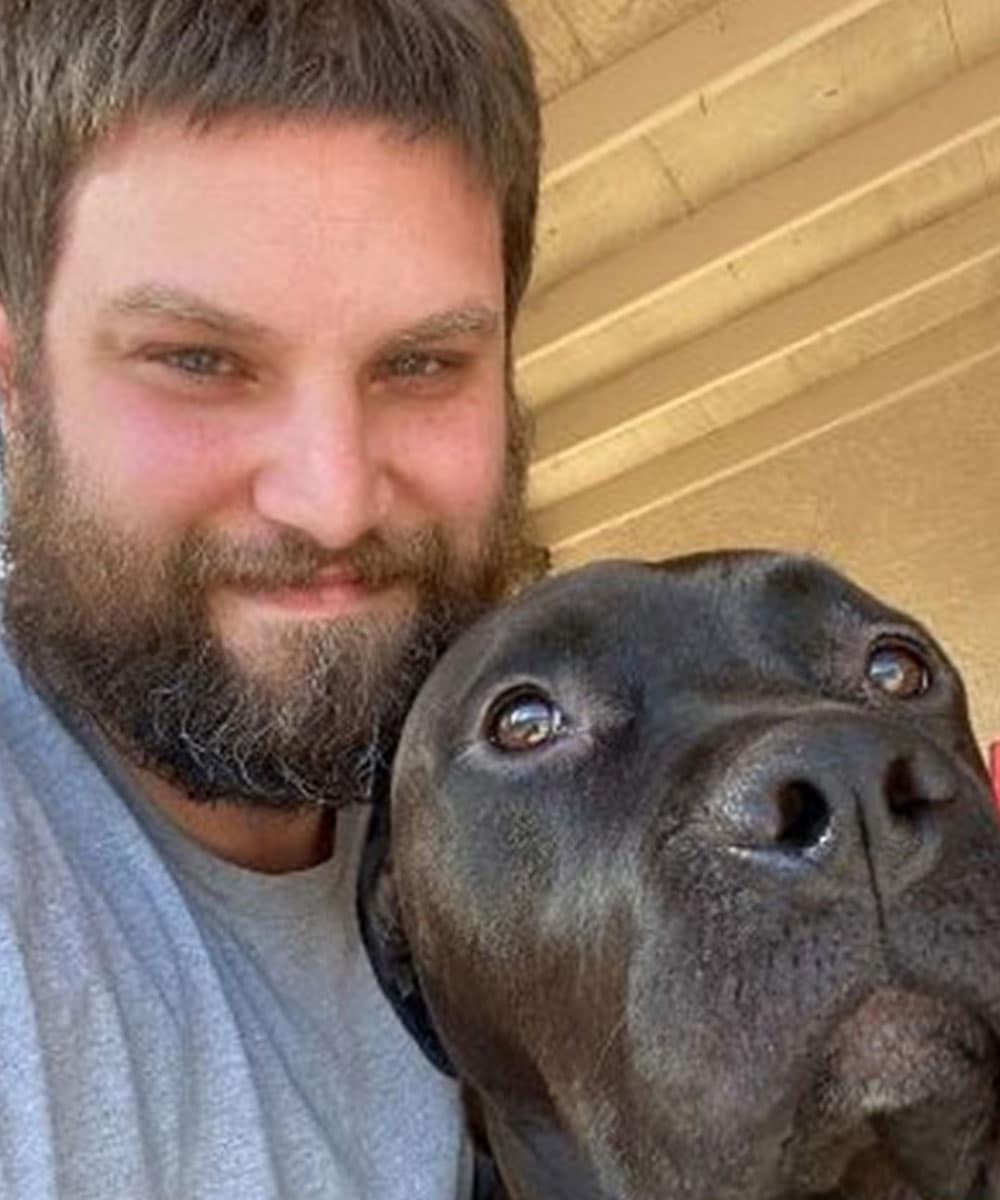 A bearded person in a light gray shirt, possibly a veterinarian, takes a selfie with a large black dog. They are both looking at the camera, surrounded by the cozy backdrop of a wooden ceiling or shelter roof.