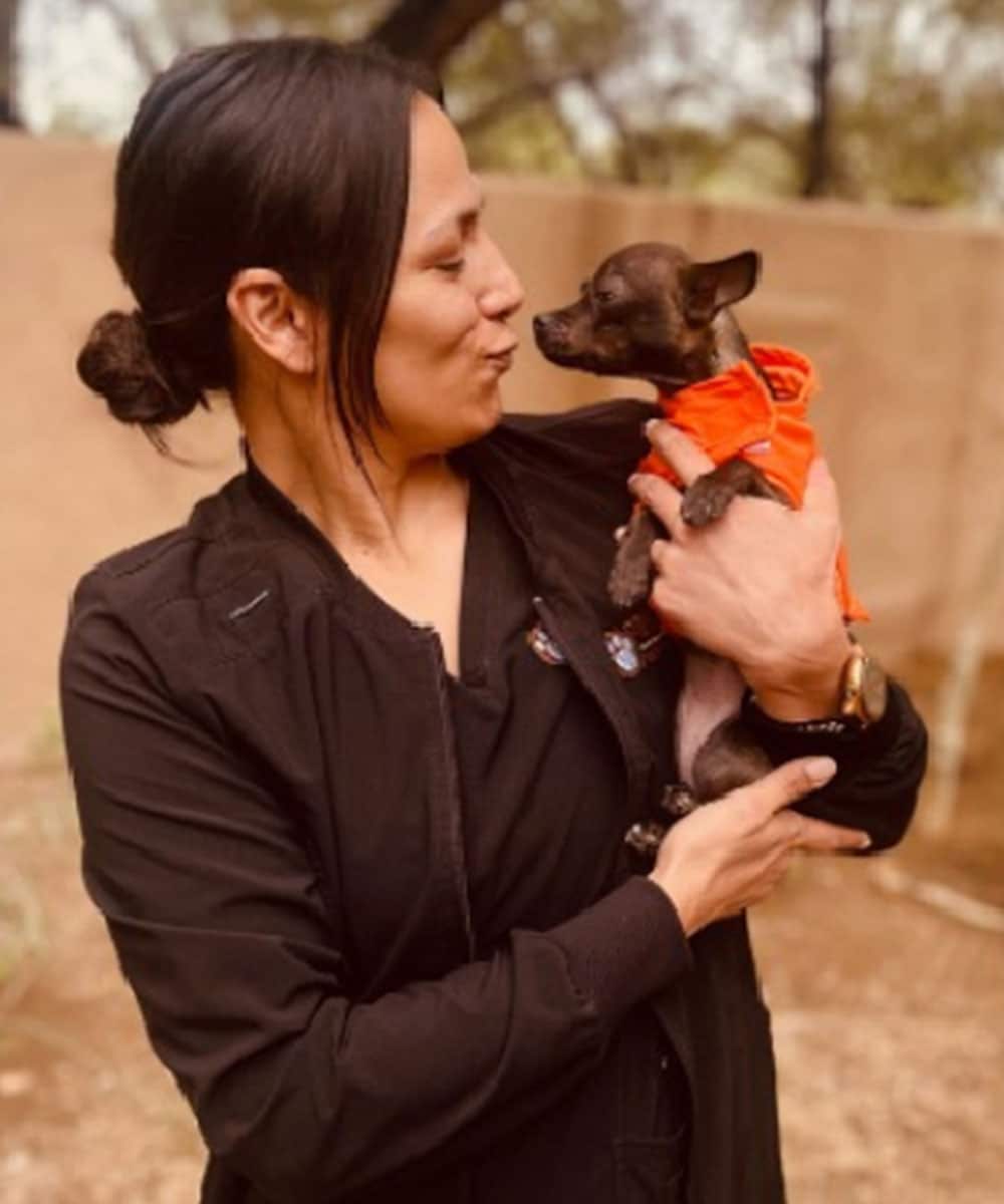 A woman in a black jacket holds a small dog wearing an orange outfit, looking like they just stepped out from a fun visit to the vet. Outdoors, she playfully purses her lips toward the pup, surrounded by blurred trees and a wall.