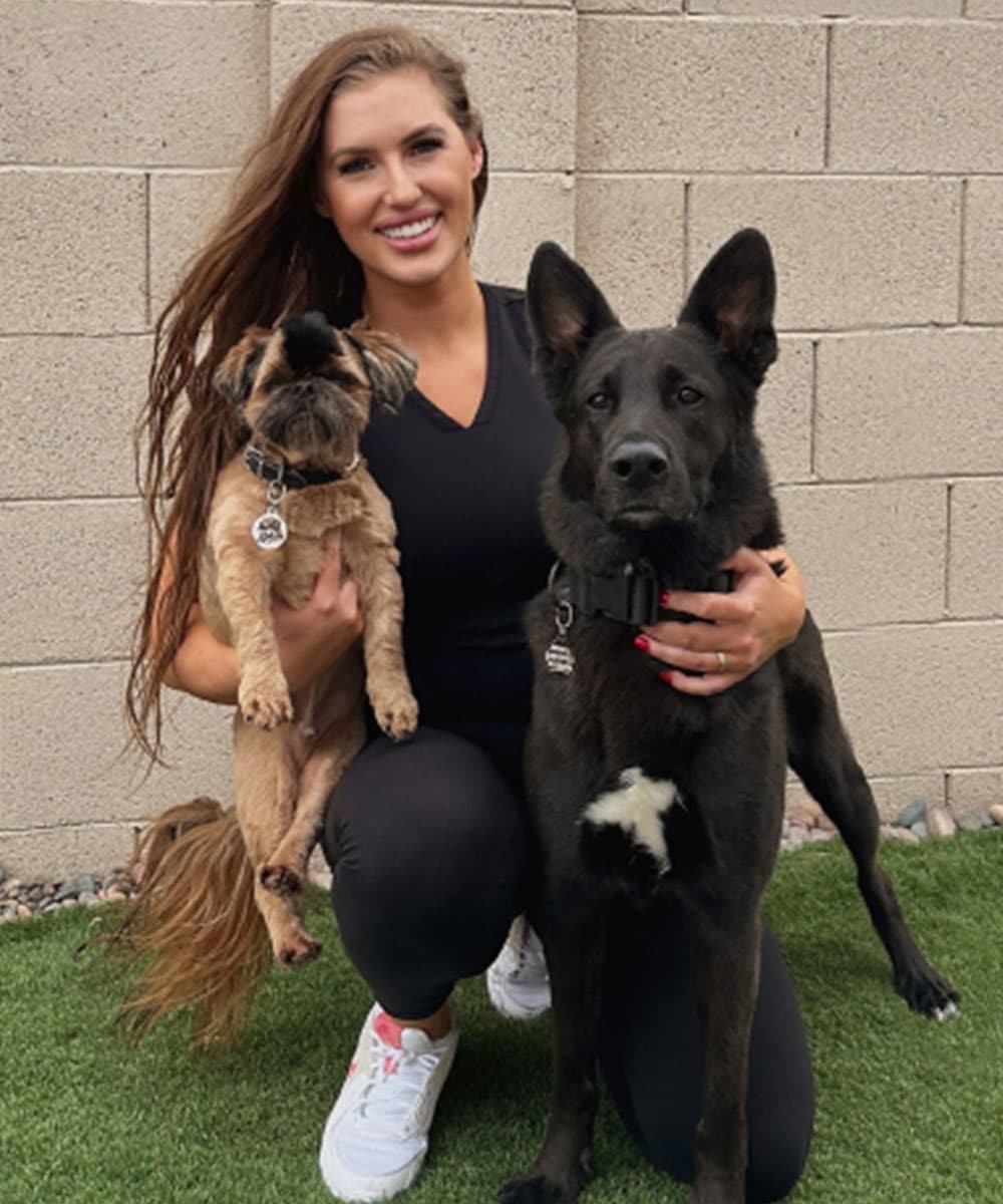 A veterinarian kneels on grass, smiling, in a black outfit and white sneakers, cradling a small brown dog. A larger black dog stands loyally beside them against a backdrop of a brick wall.