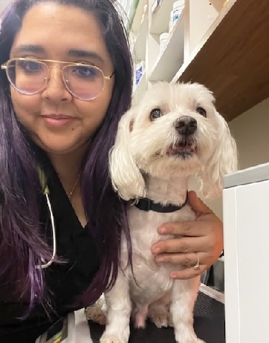 A person with long dark hair and glasses, who might just be a vet, smiles at the camera while holding a small white dog. The dog has fluffy fur, wears a black collar, and looks content. They're in an indoor setting with shelves in the background.