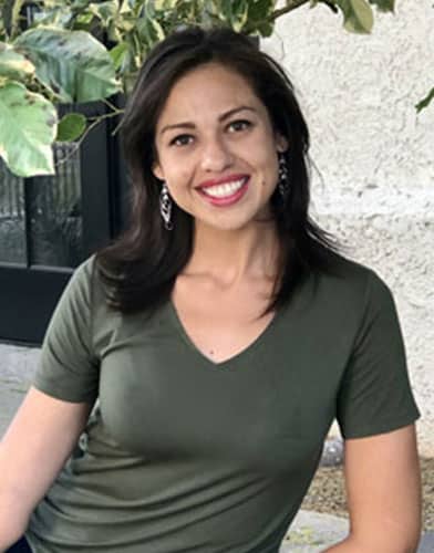 A woman with dark hair and a green shirt sits smiling in front of a plant, embodying the warmth and compassion of a dedicated vet.