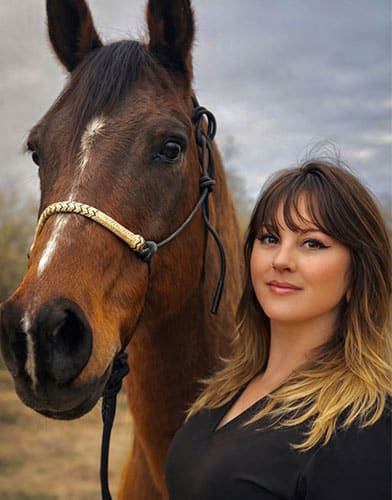 A woman with shoulder-length brown and blonde hair stands next to a brown horse with a white mark on its face. Both are outdoors with a cloudy sky in the background.