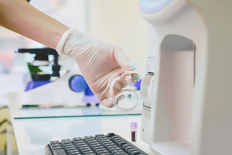 A gloved hand inserts a sample into a laboratory machine, evoking the precision of a veterinary clinic. A microscope and a small vial with a purple cap are on the table, while a computer keyboard sits in the foreground, highlighting this meticulous setting where vets unravel scientific mysteries.
