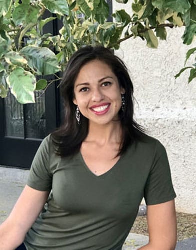A woman with dark hair, wearing a green V-neck shirt and earrings, smiles while sitting outdoors. A dedicated veterinarian, she enjoys a moment of peace under a leafy tree, with a light-colored wall and black-framed glass doors providing an elegant backdrop.