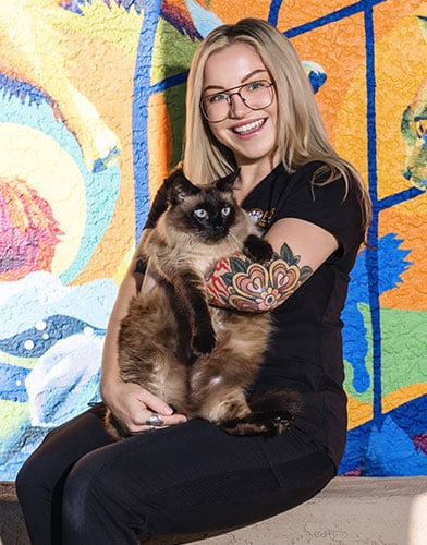 A smiling woman with glasses and a colorful tattoo on her arm holds a Siamese cat. They are sitting in front of a vibrant, colorful mural with abstract shapes and patterns.
