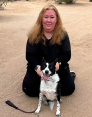 A person with long hair is kneeling on sandy ground, holding a black and white dog with a leash. This veterinarian seems to be enjoying the calmness of the outdoor setting, bonding closely with their furry companion.