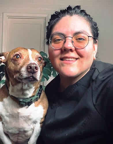 A veterinarian with glasses and dark hair smiles warmly while holding a brown and white dog. The person is wearing a black shirt, and the dog sports a green collar. In the background, there's a door framed by lush green leaves.