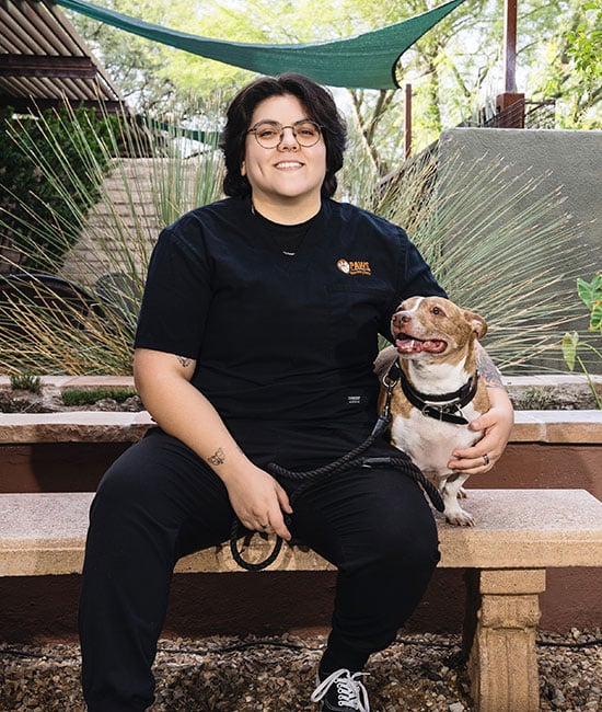 Person with glasses and short dark hair sits on a bench, smiling and holding a happy dog on a leash. As a veterinarian, they enjoy their downtime outdoors, surrounded by plants and greenery.