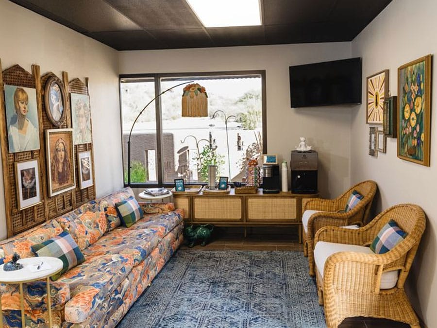 A cozy living room with a floral-patterned sofa and two wicker chairs, resembling the welcoming atmosphere of a vet's waiting area. The walls are adorned with various paintings. A large window provides natural light, and a TV is mounted in the corner above a console with a coffee maker. A rug covers the floor.