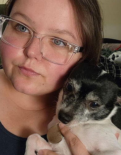 Person with clear glasses, possibly a veterinarian, holding a small black and white dog. The person has brown hair and is wearing a dark top. The background is blurred, featuring checkered fabric.