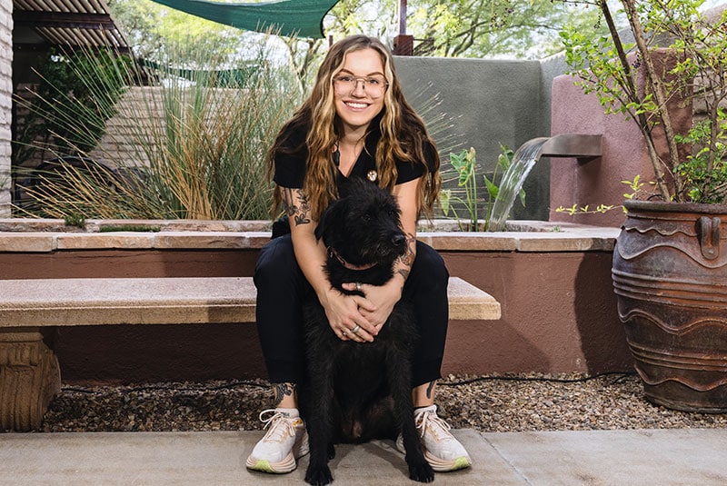 A smiling woman with long hair and glasses, who is a veterinarian, sits on an outdoor bench holding a fluffy black dog. They enjoy the garden setting together, surrounded by lush plants, gravel pathways, and a soothing water feature in the background.