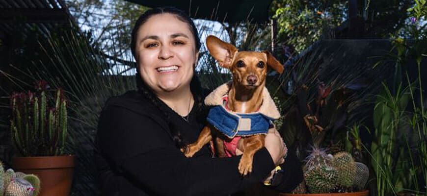 A smiling person with dark hair in braids holds a small brown dog wearing a denim jacket. They are outdoors, surrounded by green plants and cacti in pots.