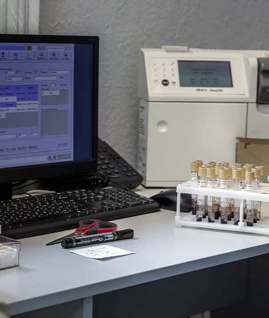 A lab desk with a computer displaying veterinary data, a marker, and a stand holding test tubes filled with samples. A printer is partially visible in the background.