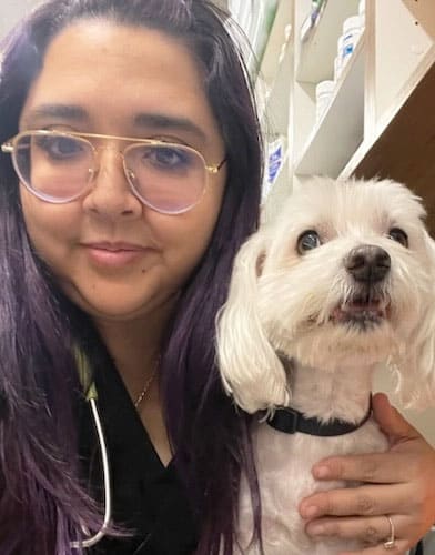 A person with long dark hair and glasses smiles while holding a small white dog. The veterinarian, with a stethoscope around their neck, stands in what appears to be a veterinary setting, as shelves filled with containers are visible in the background.