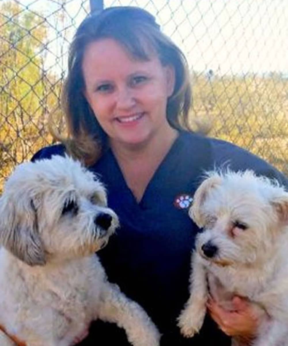 A person smiling and holding two fluffy white dogs in an outdoor setting, with a chain-link fence and blurred natural background. The veterinarian, wearing a dark top with a paw print design, exudes warmth and care.