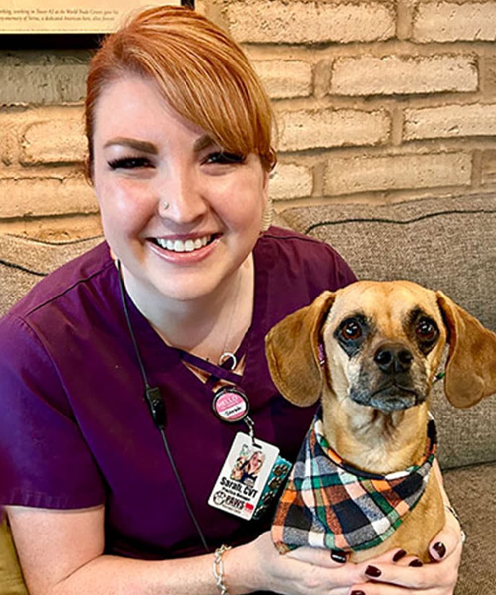 A smiling person with red hair, wearing purple scrubs, sits next to a small brown dog wearing a plaid bandana. As a dedicated veterinarian, they share a bond in the cozy room with a brick wall backdrop.