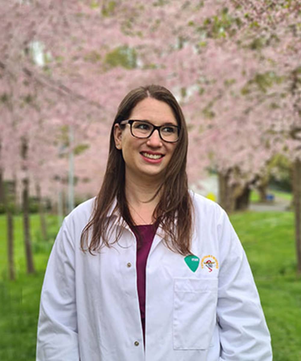 A woman with long brown hair and glasses wearing a white lab coat stands outside in front of blooming cherry blossom trees, smiling and looking to the side. As a dedicated veterinarian, she takes a moment to enjoy nature's beauty.