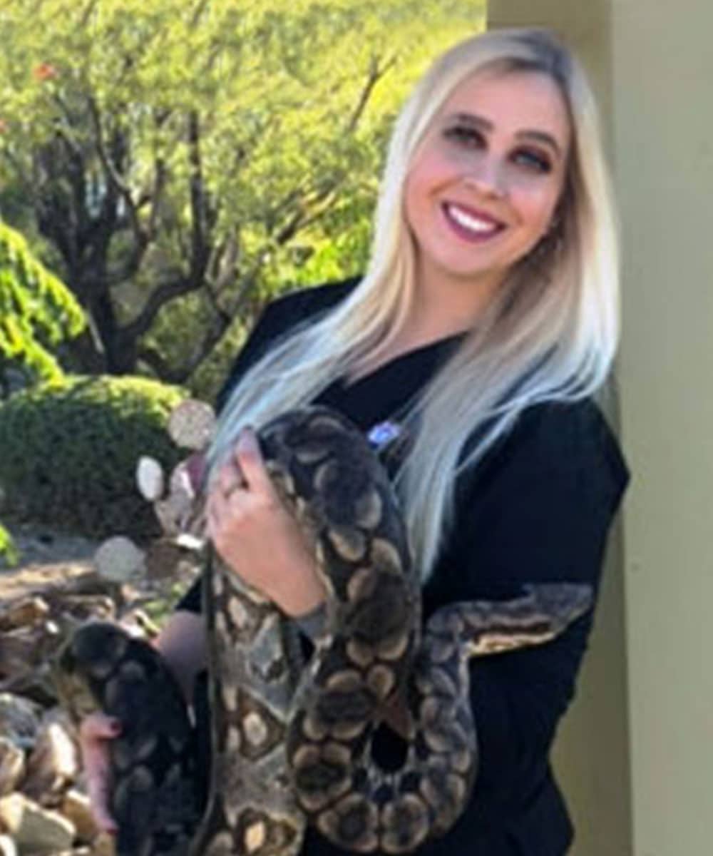 A woman with long blonde hair, dressed in a black outfit, smiles as she holds a large snake with a brown and tan pattern. As a veterinarian, she's at ease amidst the green foliage and sunny outdoor setting, showcasing her love for all creatures great and small.