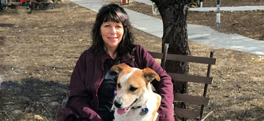 A woman sits on a wooden chair outdoors, smiling with a brown and white dog on her lap. As a caring veterinarian, she enjoys these peaceful moments. The background features a tree and a concrete path surrounded by dirt.