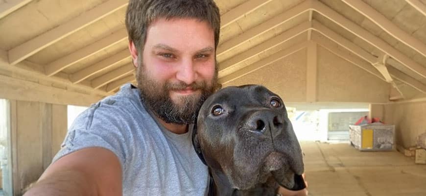 A bearded man, perhaps a vet, smiles for a selfie with his black dog in a spacious, unfinished building. Both look warmly at the camera, set against a backdrop of wooden beams and scattered construction materials.