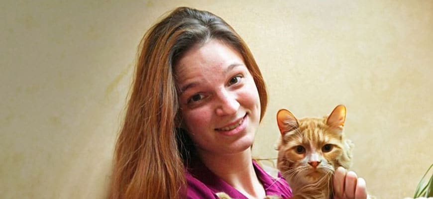 A smiling veterinarian with long brown hair holds an orange tabby cat. The background is a plain, light-colored wall.