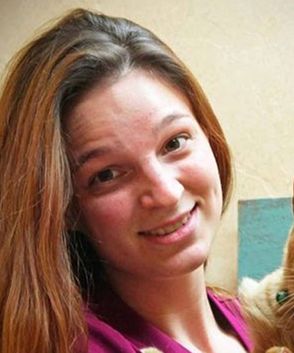 A person with long, reddish-brown hair is smiling, perhaps a veterinarian in their off-hours. Indoors, they wear a dark pink top, with part of their face partially obscured by a light brown object. The background features a light-colored wall with hints of blue.