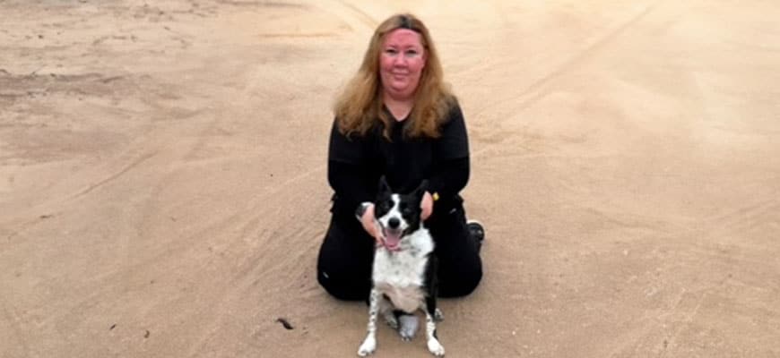A woman with long hair, wearing a black outfit, kneels on a sandy beach beside her black and white dog, who she always calls her best patient. The background is a vast, empty stretch of sand.
