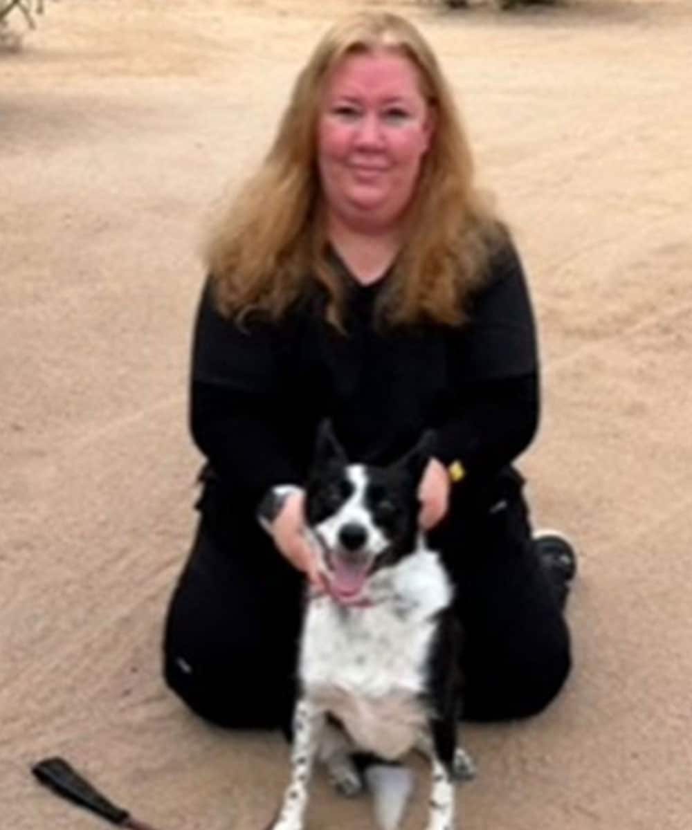 A person with long hair kneels on the sand, smiling alongside a black and white dog, both gazing warmly at the camera. The person, dressed in black, exudes the gentle care of a veterinarian. A brush rests nearby on the ground.