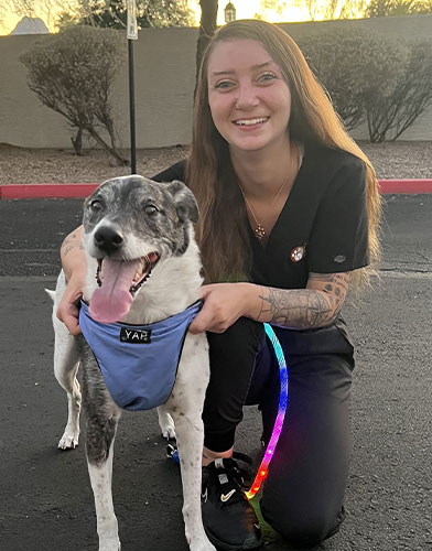 A woman in black scrubs kneels next to a happy, black-and-white dog wearing a blue harness and a rainbow-colored leash, both smiling outdoors on a paved surface.