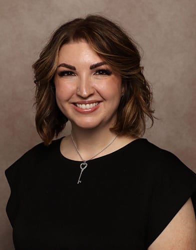 A woman with shoulder-length brown hair, wearing a black top and a silver key-shaped necklace, smiles at the camera against a soft brown background.
