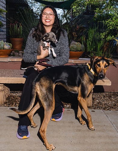 A smiling person sits on a bench outside, holding a small puppy, while a large black and brown dog stands in front of them, looking toward the camera. Potted plants are visible in the background.