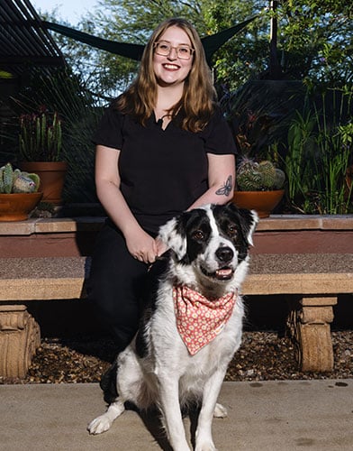 A woman with long, light brown hair and glasses sits on a bench outside, smiling. A black and white dog wearing a red patterned bandana sits in front of her. Lush plants and trees are in the background.