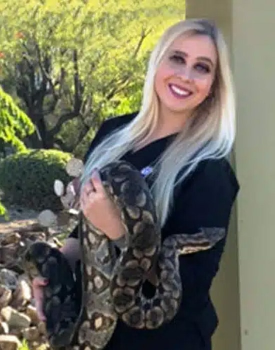 A smiling woman with long blonde hair holds a large snake in her arms while standing outdoors near greenery and rocks.
