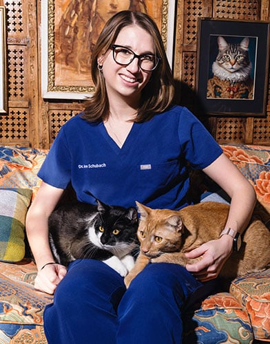 A woman in blue scrubs, sitting on a patterned couch, smiles while holding a black and white cat and an orange cat. Framed cat portraits hang on the wooden wall behind her.