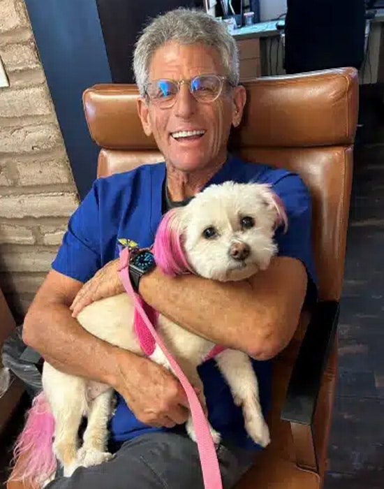 An older man with gray hair and glasses smiles while sitting in a brown chair, holding a small white dog with pink ears and a pink leash in his arms.