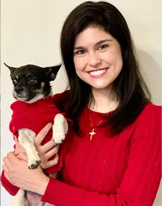 A woman with long dark hair, wearing a red sweater and a gold cross necklace, smiles while holding a small black and white dog dressed in a matching red sweater.