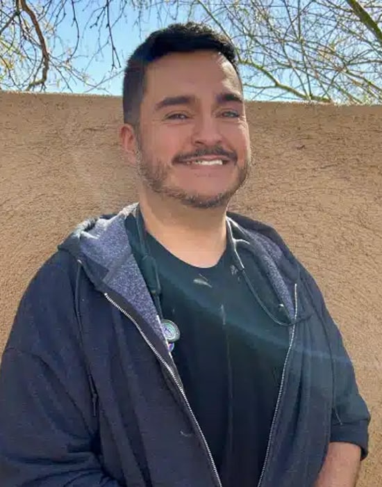 A person with short dark hair and facial hair smiles while standing outdoors in front of a tan wall, wearing a black shirt and dark zip-up hoodie. Leafless branches and a blue sky are visible in the background.