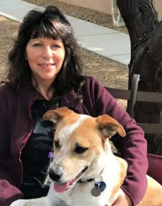 A woman with long dark hair sits on a bench outdoors, smiling, with a brown and white dog on her lap. The dog is looking slightly to the side with its tongue out. Trees and a sidewalk are in the background.