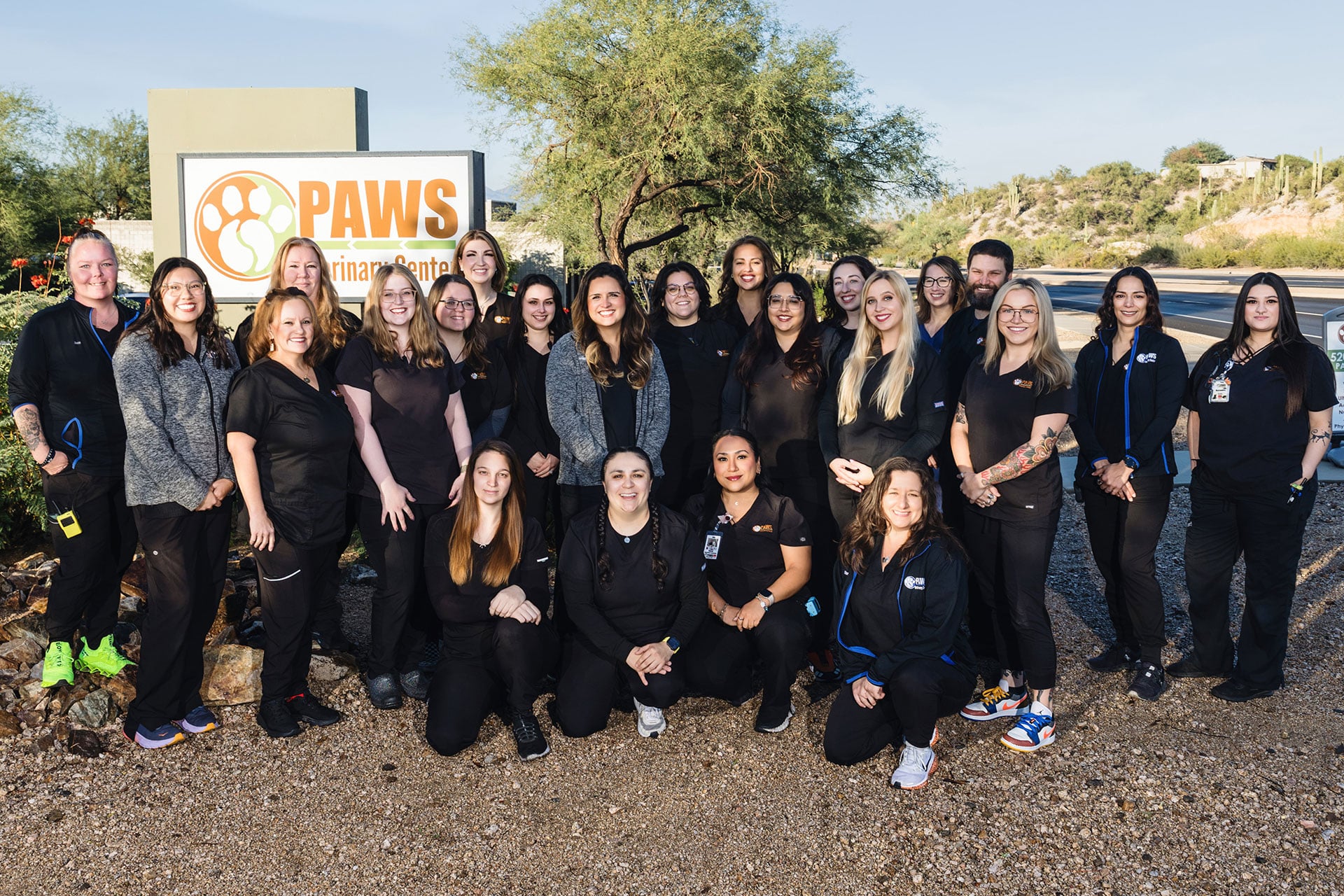 A group of about 23 people, mostly women, stand and kneel together outdoors in front of a sign reading “PAWS.” They are smiling, dressed in black attire, with trees and a road in the background.