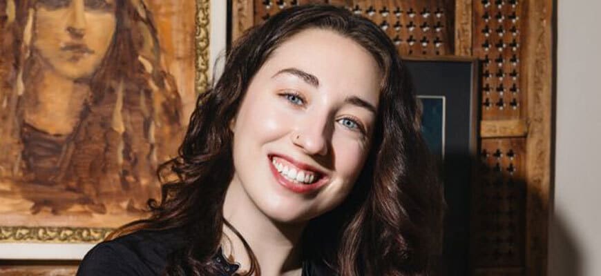 A woman with long brown hair and blue eyes, smiling at the camera, stands indoors in front of a framed painting and a wooden panel with a lattice design.
