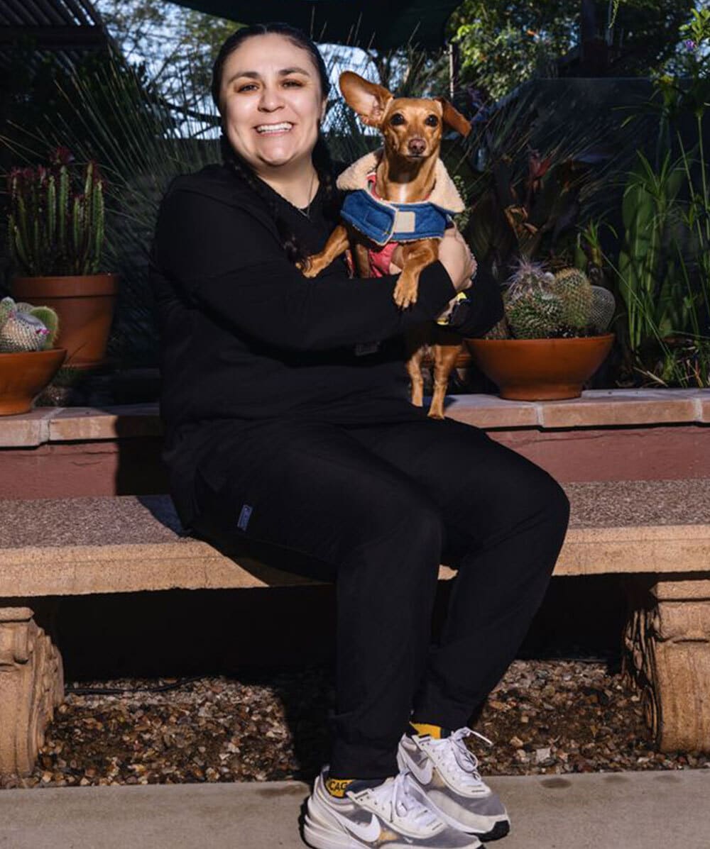 A smiling person in black clothing sits on a bench outside, holding a small brown dog in a blue harness and tan jacket. There are potted plants and cacti in the background.