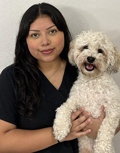 A woman with long dark hair and a black shirt holds a small, fluffy white dog. Both are looking at the camera and the dog has its mouth open, appearing to smile. The background is a plain light-colored wall.